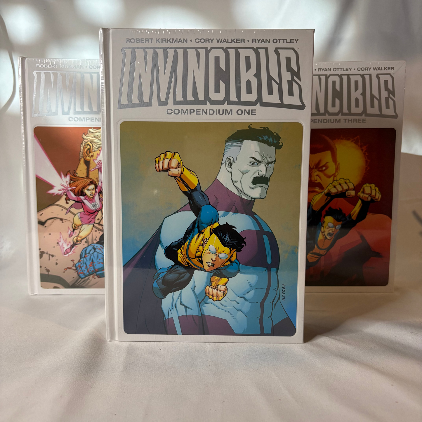 Three comic book volumes from the Invincible Compendium One series displayed on a white fabric surface under dappled light. The center volume is upright and clearly shows the title 'INVINCIBLE COMPENDIUM ONE' with cover art of Invincible and a large robot face.