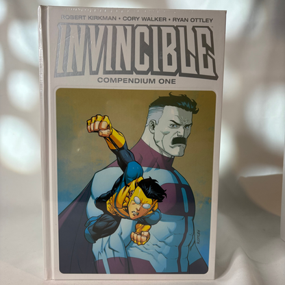 Invincible Compendium One graphic novel standing upright on a white fabric surface with soft shadow overlays. The cover shows the title 'INVINCIBLE' and illustrated characters in blue, yellow, black, and purple tones.